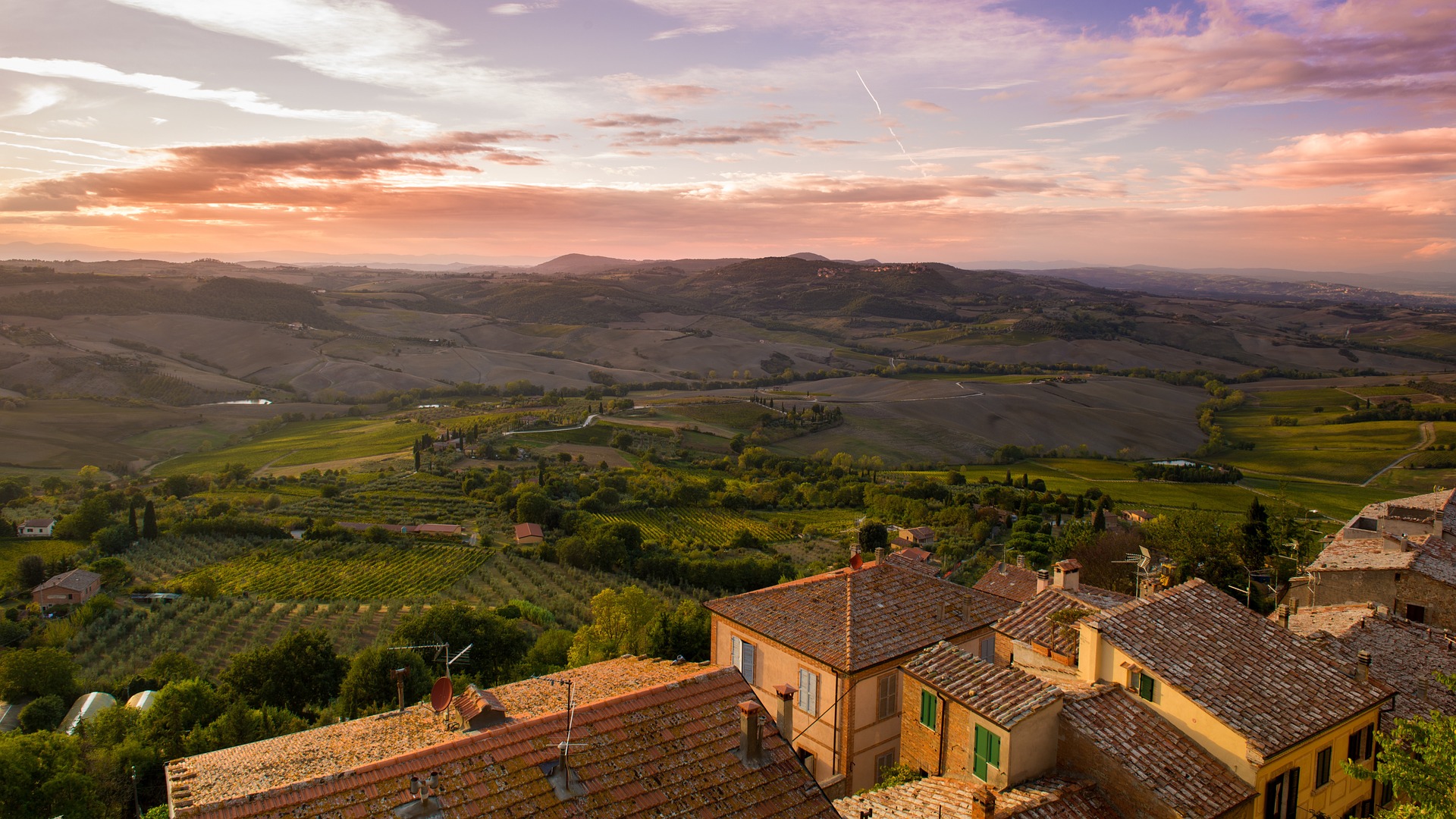 Tuscany Italy at sunset, overlooking a beautiful hillside with pink and purple sky, green trees grass, and buildings in the foreground